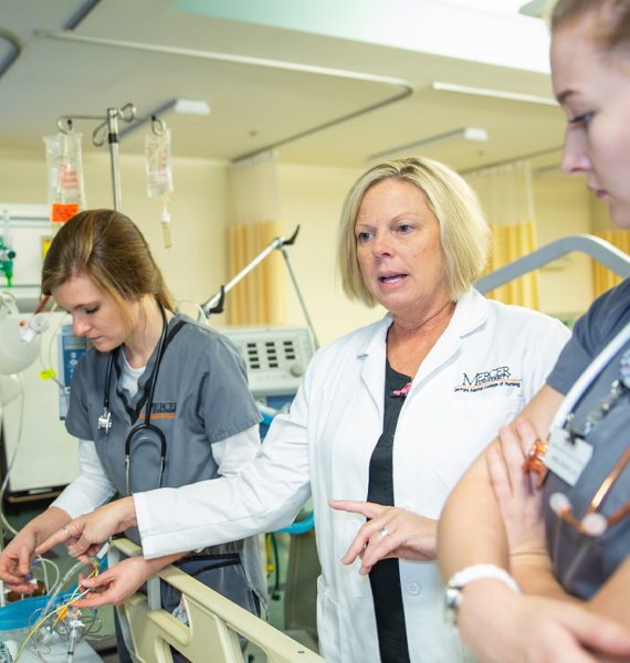 Two nursing students in scrubs with stethoscopes around their necks work with a faculty member in a white coat