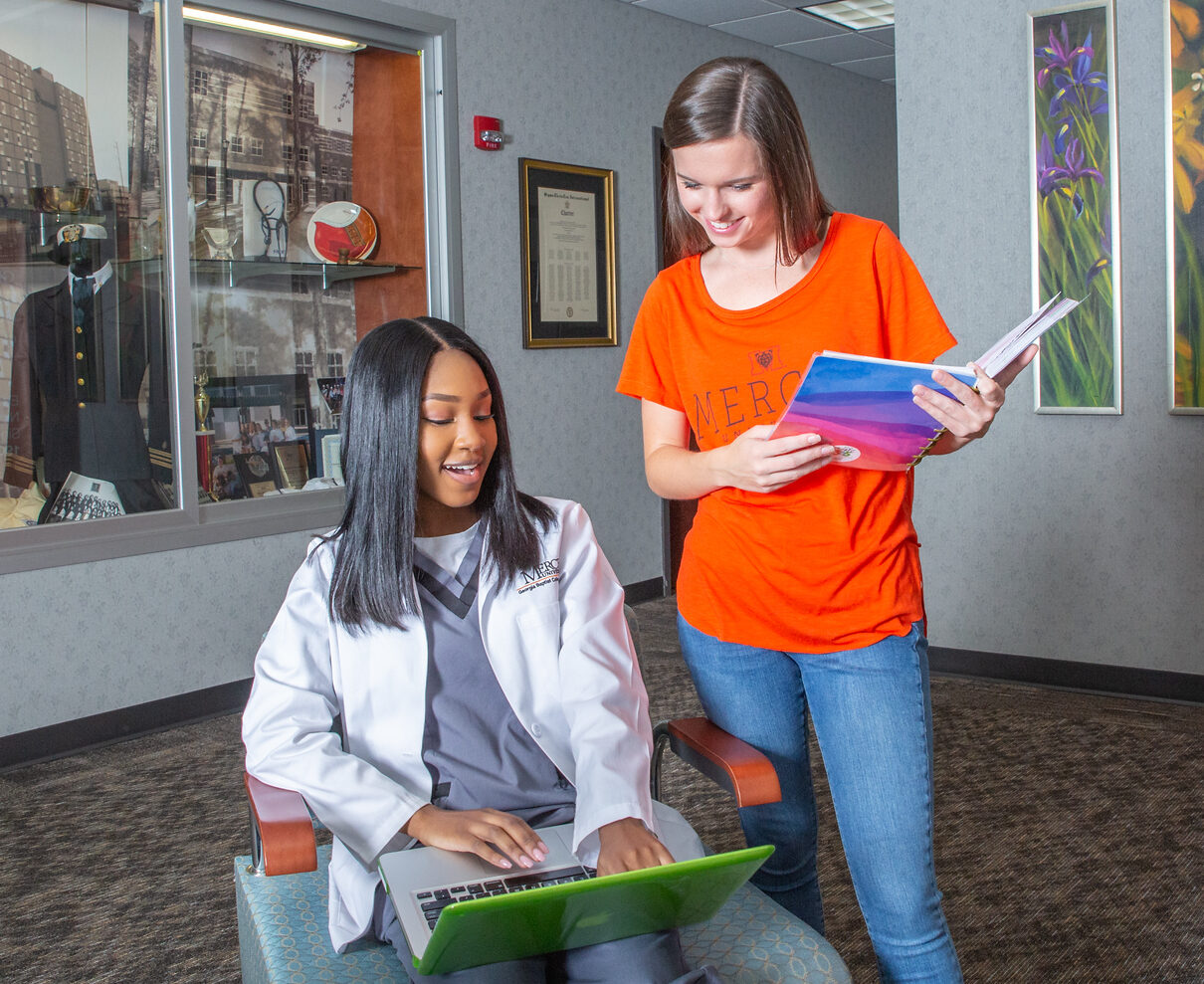 Two nursing students, one in scrubs in chair on laptop, conversing with student in Mercer shirt standing beside sitting student