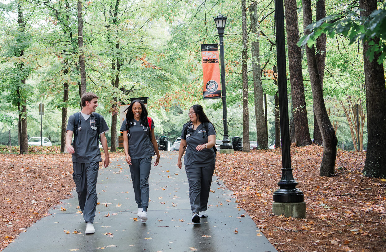 three Mercer BSN students walking in scrubs on campus, green trees in background